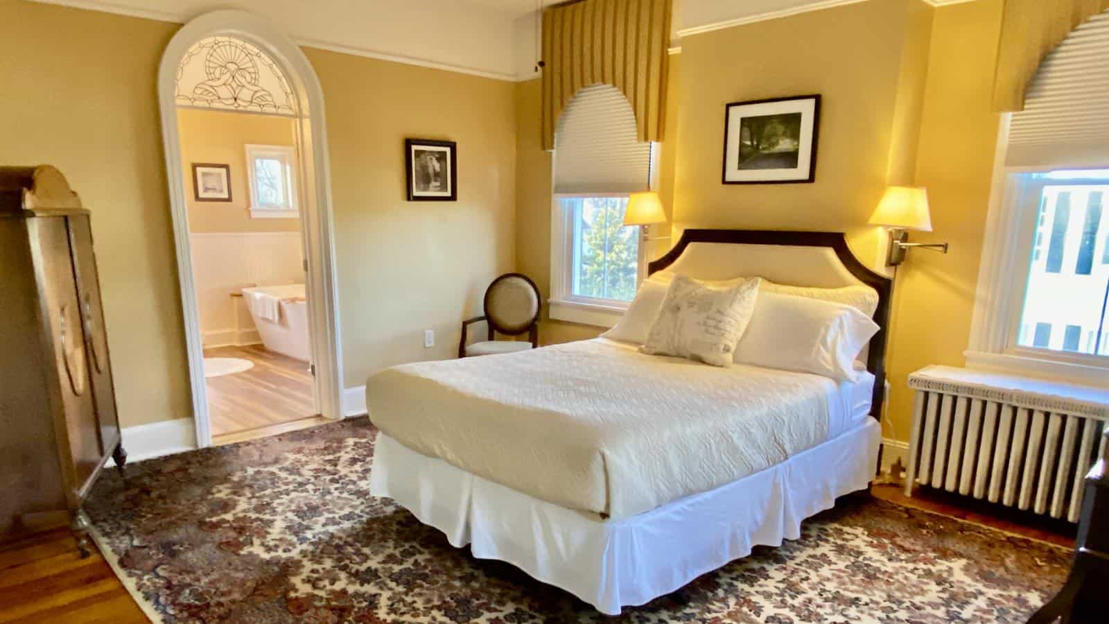 Bedroom with yellow walls, hardwood flooring, white bedding, dark wooden armoire, antique chair, and view into bathroom