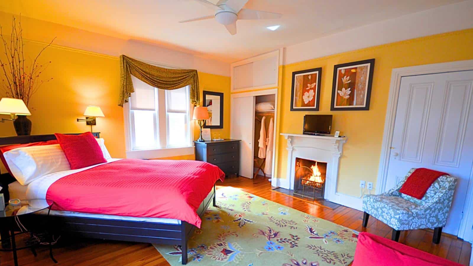 Bedroom with yellow walls, hardwood flooring, dark wooden bed, red bedding, fireplace, dresser, and upholstered armchair