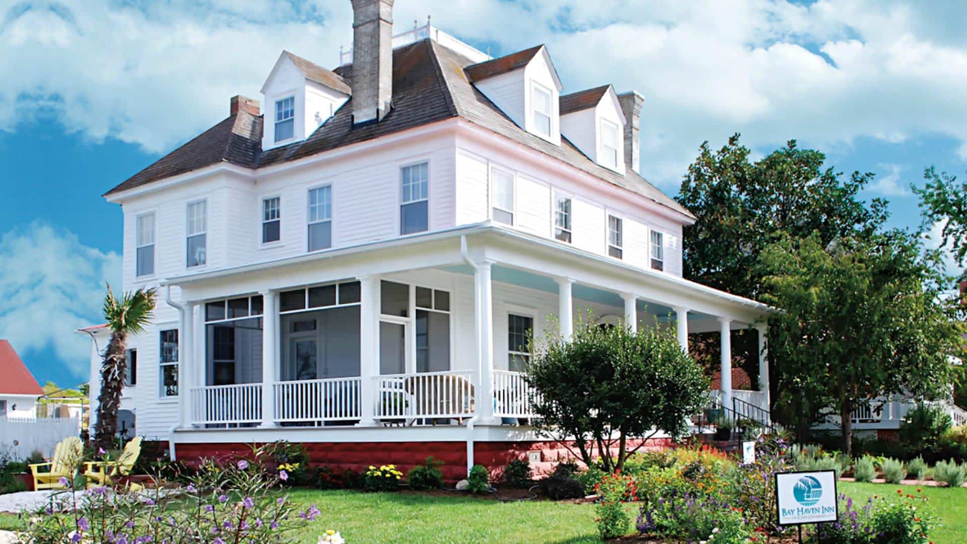 Exterior view of property painted all white with wrap around porch surrounded by green grass, shrubs, flowers and trees