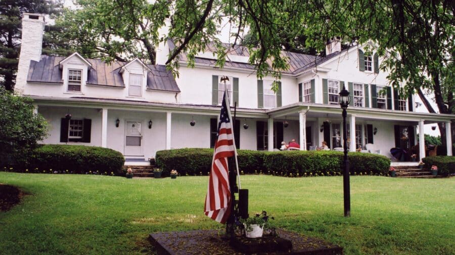 Exterior view of property painted white with dark green shutters surrounded by green grass, shrubs, and trees