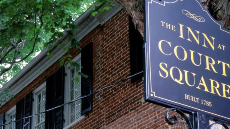 Exterior view of the property with red brick, white windows, and black shutters surrounded by green trees and a sign that says The Inn at Court Square built 1785