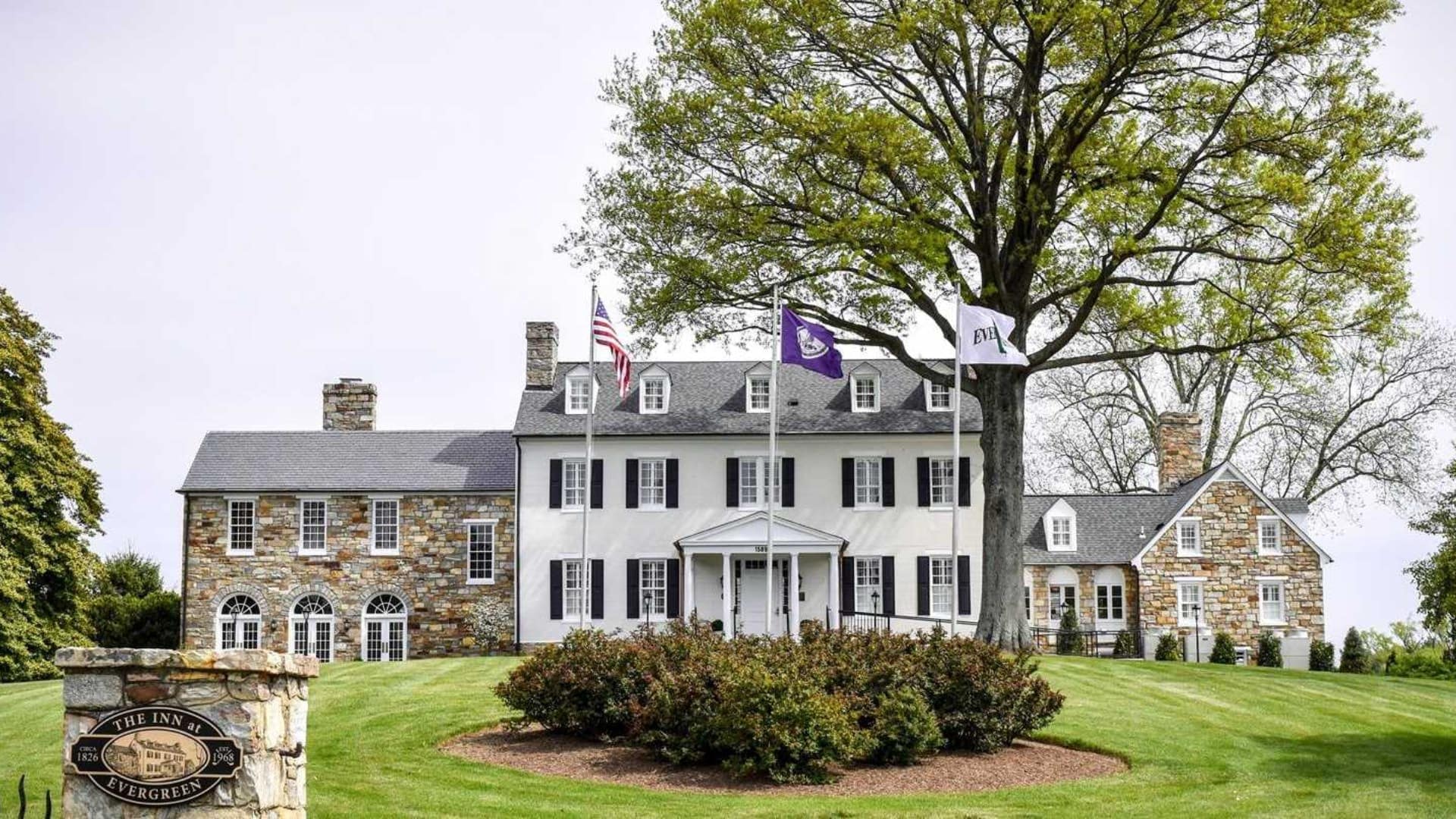 Exterior view of the property painted white with black shutters and stone brick surrounded by green grass, bushes, and trees