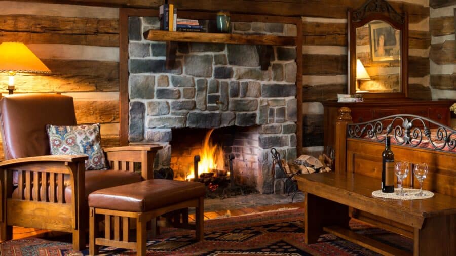 Bedroom with log cabin walls, stone fireplace, hardwood flooring, dark wooden bed and dresser, and wooden chair with brown leather cushions