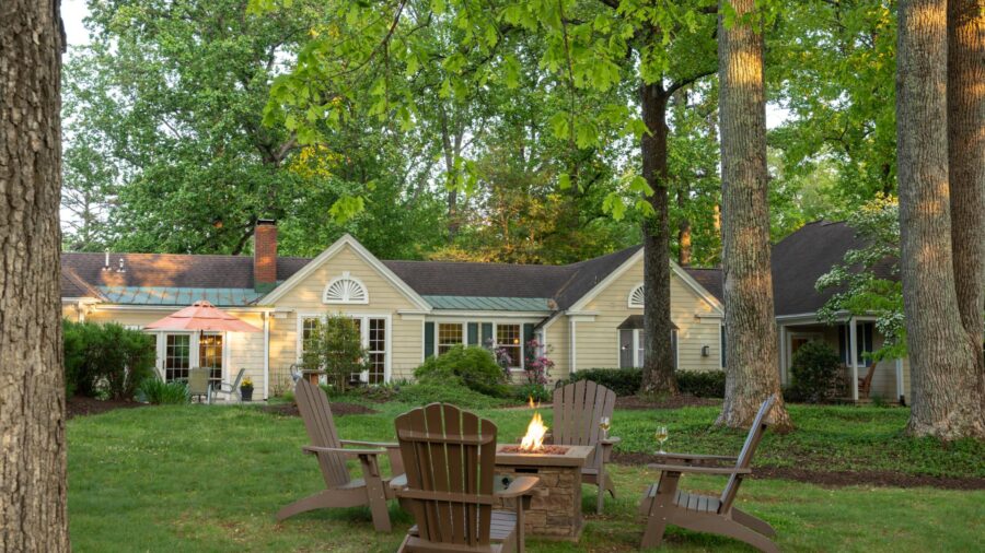 Exterior view of property painted yellow with white trim and firepit with four brown Adirondack chairs surrounded by green grass and very tall green trees