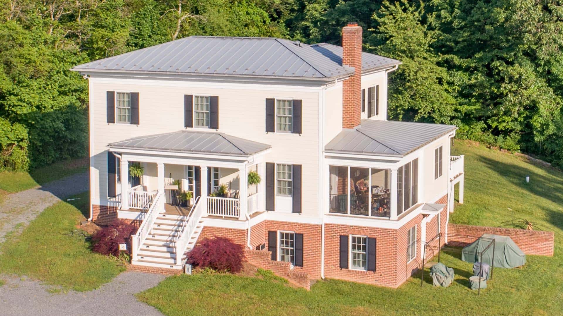 Exterior view of property painted white with black shutters and red brick for garden level of house surrounded by green grass and large green trees