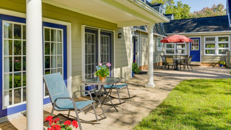 Exterior view of property painted light taupe with white trim and blue doors, paver patio with patio dining table and chairs and red umbrella and green grass