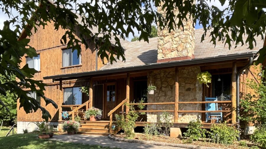 Exterior view of property with wooden siding, stone brick fireplace, and front porch surrounded by green grass, shrubs, and trees