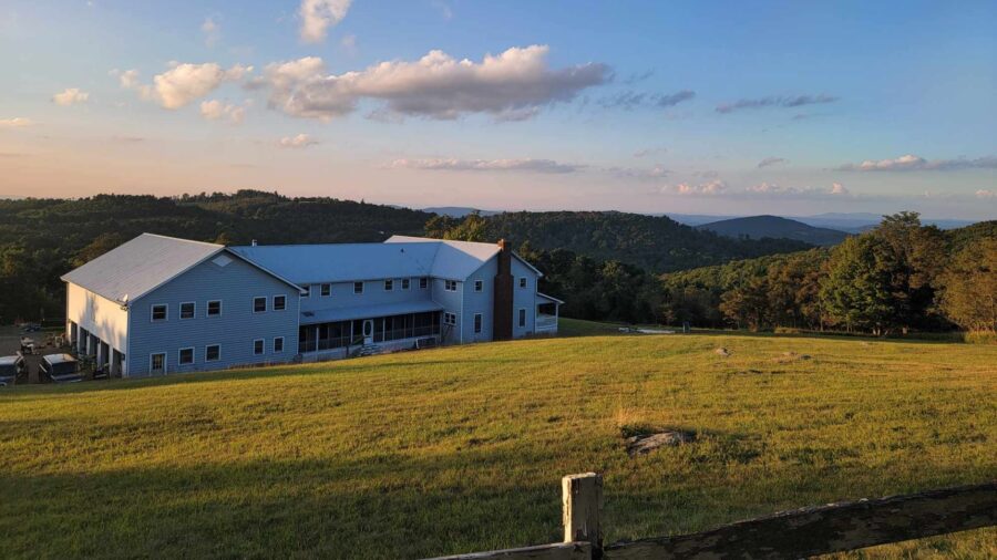 Exterior view of property painted gray with white trim surrounded by large field of grass and rolling hills with green trees in the background