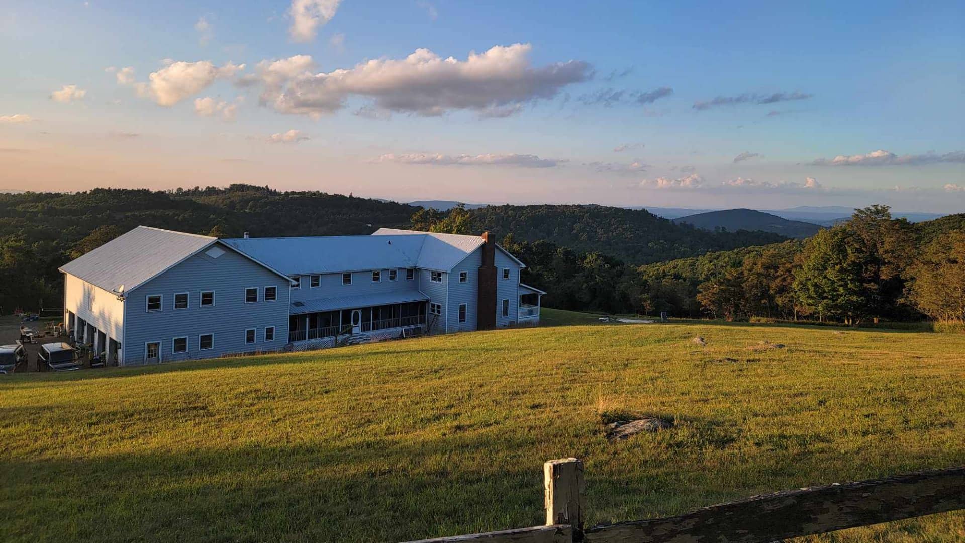 Exterior view of property painted gray with white trim surrounded by large field of grass and rolling hills with green trees in the background