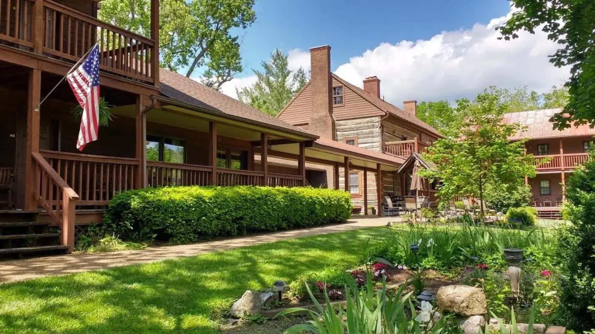 Exterior view of property with wooden siding, brown trim, large front porch, green trees, green grass, and flower gardens