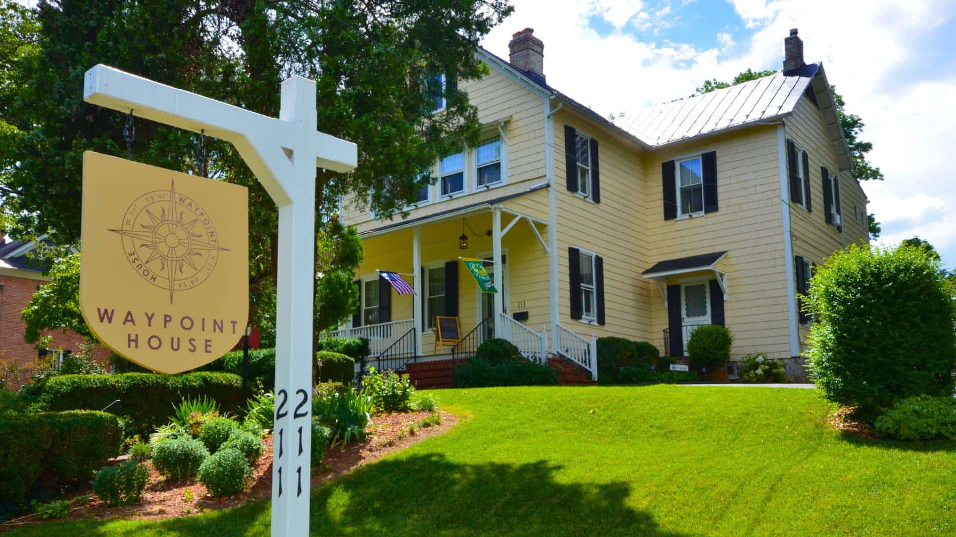 Exterior view of property painted yellow with white trim and black shutters surrounded by green grass, shrubs, gardens, and trees