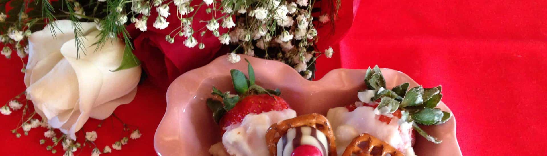 Close up view of pink heart shaped dish with white chocolate covered strawberries and a red and white rose on a red tablecloth